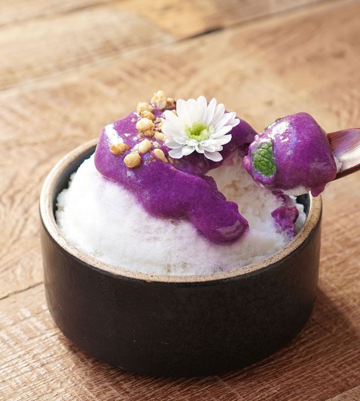 A spoon full from a bowl of shaved ice with purple paste and a white flower on top.