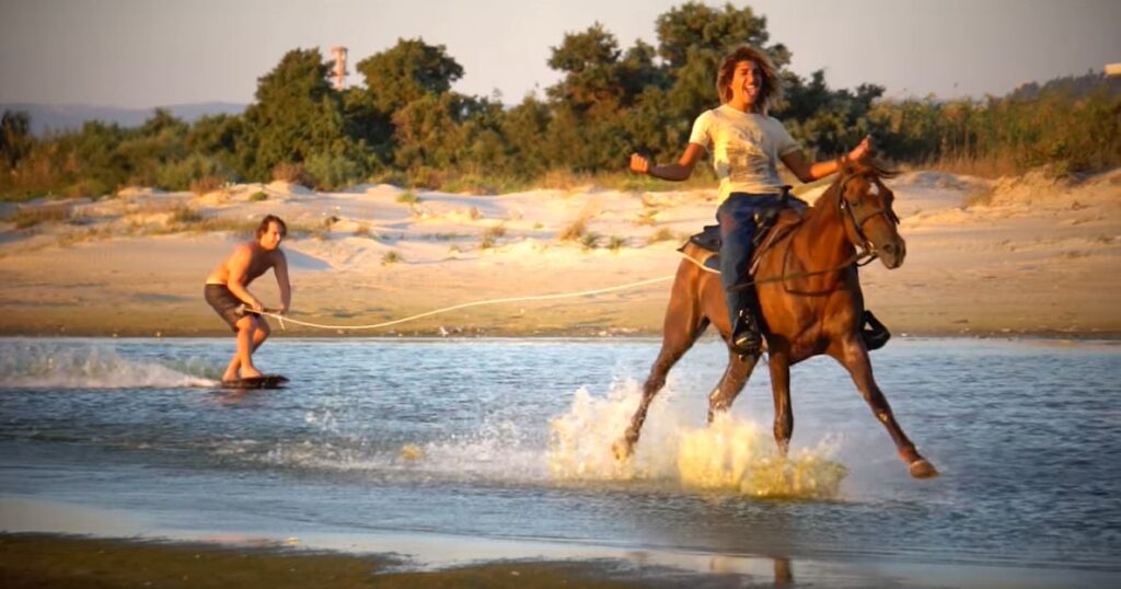 A guy riding a horse while towing another riding a surfboard.