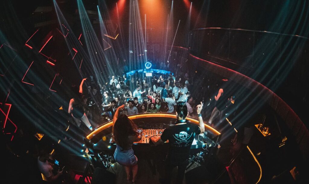 A woman and a man inside a DJ booth in front of a dancing crowd.