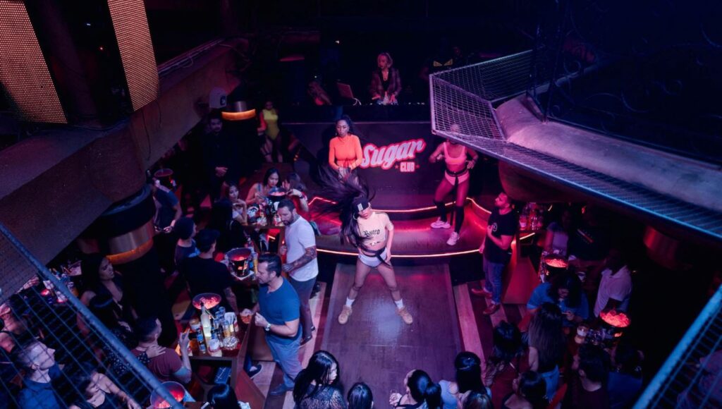 A nightclub with dancers in front of DJ booth and people drinking around.