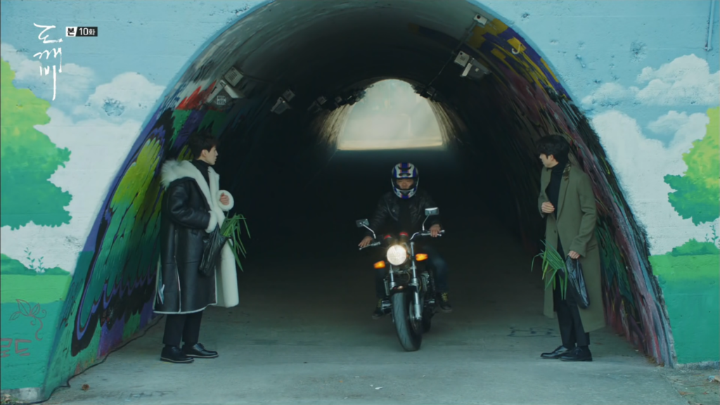 2 men watching a passing motorcycle between them in a tunnel.