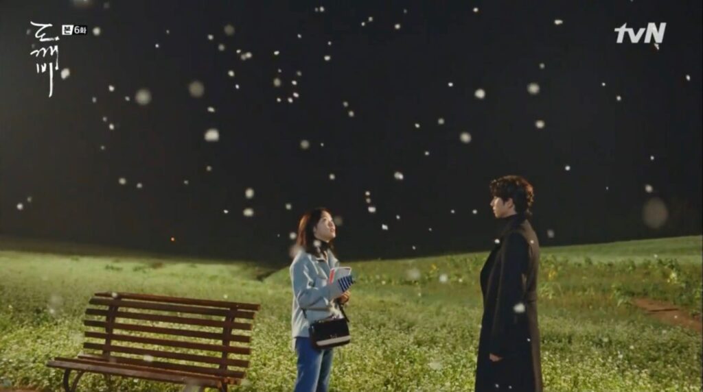 A man and a woman standing next to a bench in a green field while snowing.